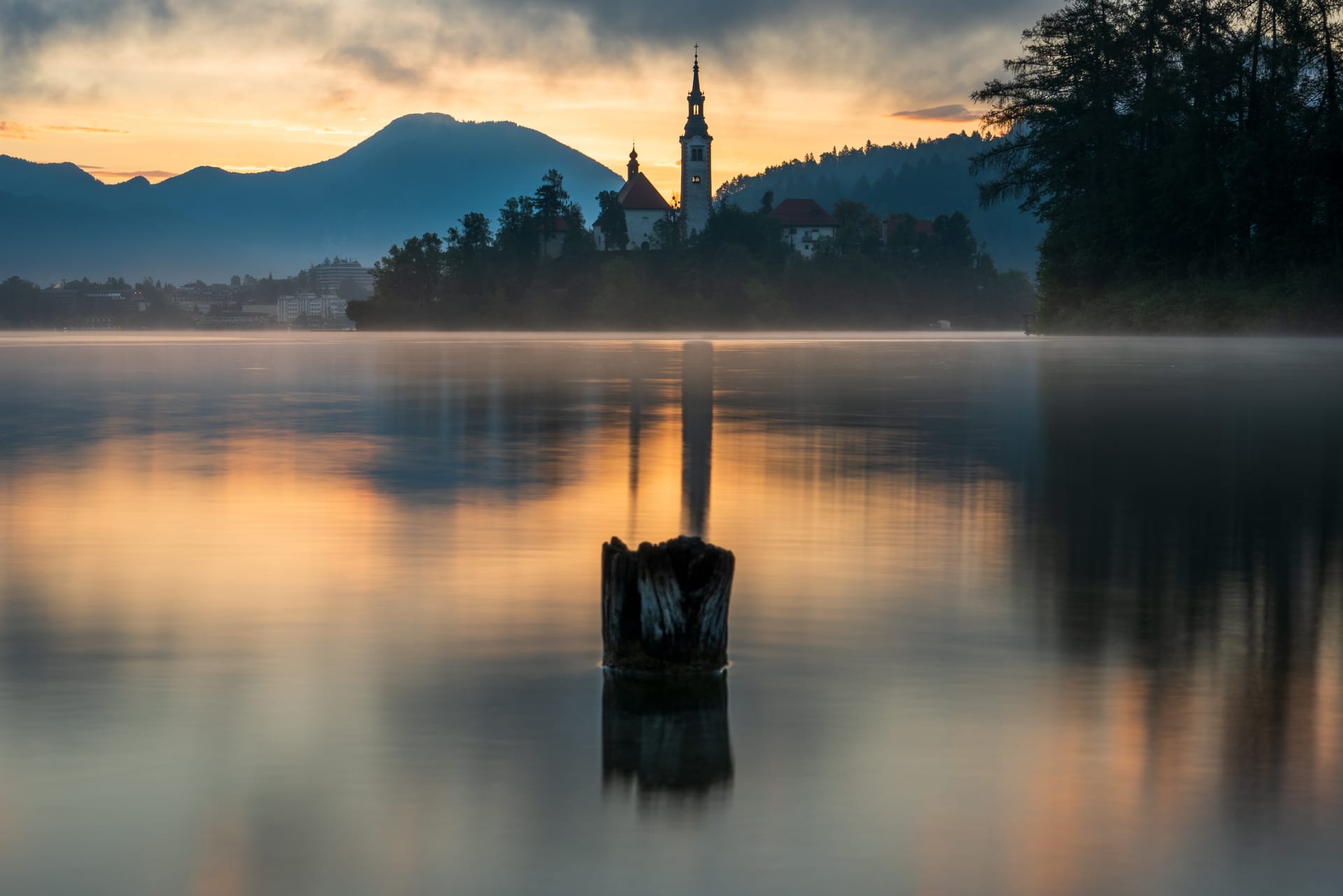 Die Wallfahrtskirche auf der Insel im Bleder See spiegelt sich im nebelverhangenen Wasser, im Vordergrund ein alter Baumstumpf, Berge im warmen Morgenlicht