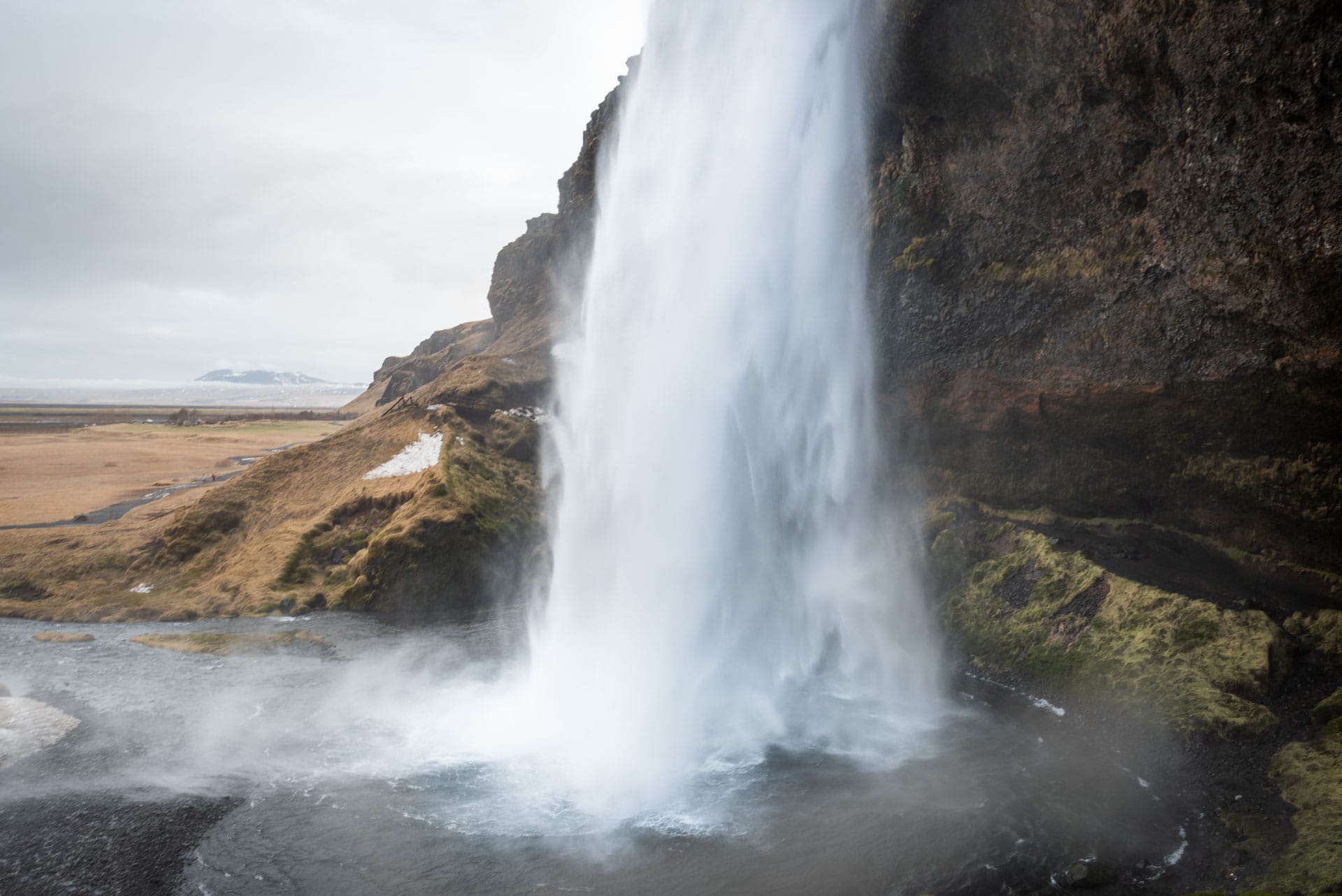 Blick aus der Höhlung hinter dem Seljalandsfoss auf den herabstürzenden Wasserstrahl, dahinter die weite Küstenebene