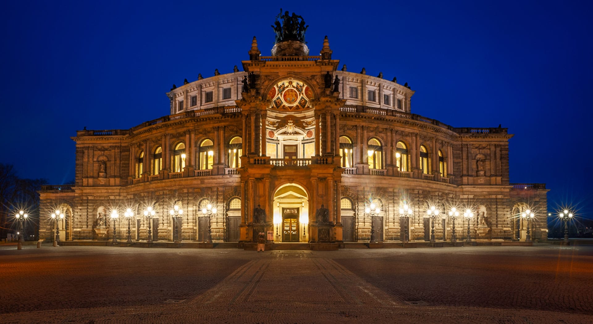 Die beleuchtete Fassade der Semperoper in Dresden vor tiefblauem Abendhimmel, das nasse Pflaster des Theaterplatzes spiegelt das Licht